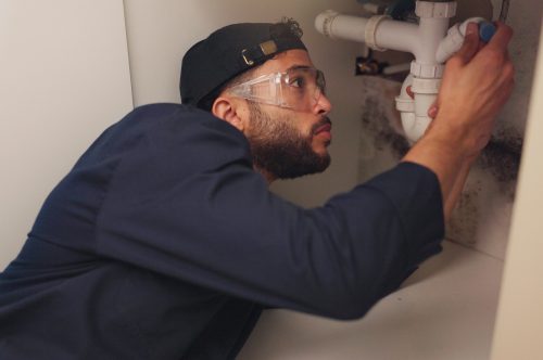 Plumber, man and pipeline repair in kitchen with tools for home maintenance and fixing leak by sink